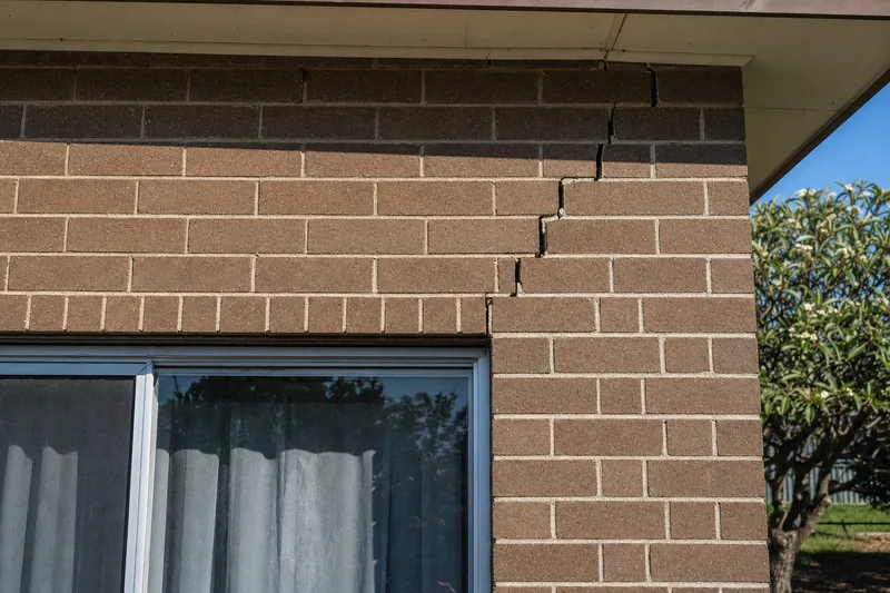 A stair-step crack running through the mortar joints of a brown face-brick wall above the head of a sliding window — crack repair, Brisbane. Illustrative example.